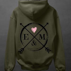 Può includere: Felpa con cappuccio verde oliva con grafica sul retro. La grafica presenta un cuore rosa all'interno di un cerchio nero, le lettere "E" e "M", frecce incrociate e una e commerciale.