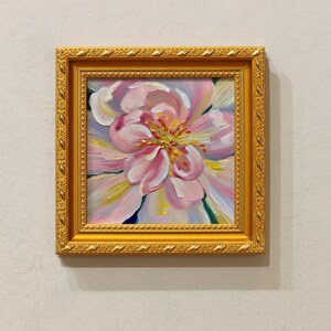 May include: A framed floral oil painting. The artwork features a close-up of a pink and white flower with yellow and green accents. The painting is set in a gold-colored ornate frame, creating a classic and elegant aesthetic.