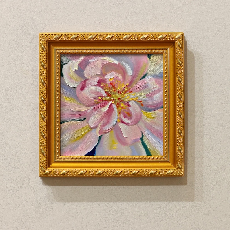 May include: A framed floral oil painting. The artwork features a close-up of a pink and white flower with yellow and green accents. The painting is set in a gold-colored ornate frame, creating a classic and elegant aesthetic.