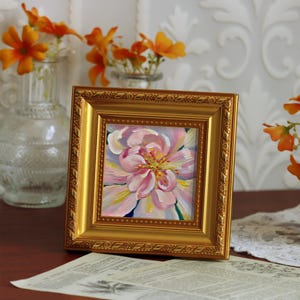 May include: A framed floral painting with a gold ornate frame. The painting depicts a close-up of a pink and yellow flower. The painting is displayed on a dark wooden surface with a vintage paper and a vase of orange flowers in the background.