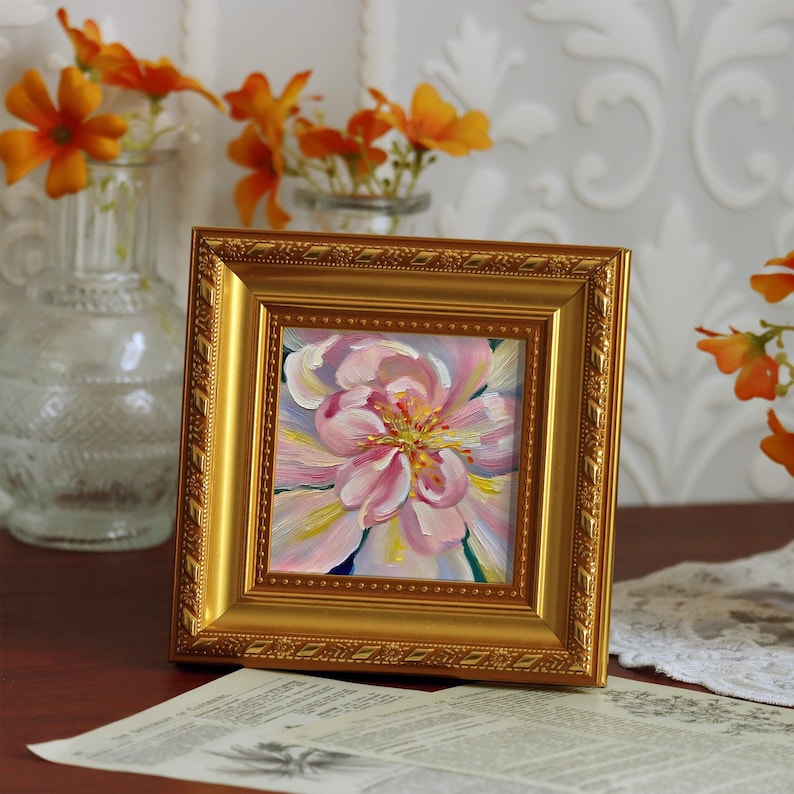 May include: A framed floral painting with a gold ornate frame. The painting depicts a close-up of a pink and yellow flower. The painting is displayed on a dark wooden surface with a vintage paper and a vase of orange flowers in the background.