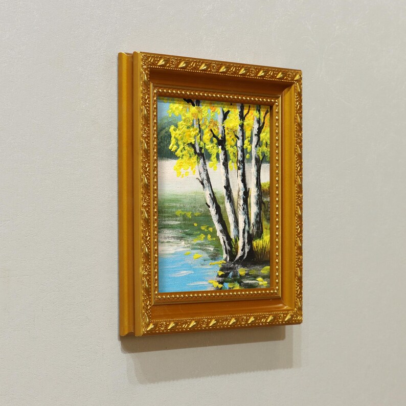 Op de afbeelding: Een ingelijst landschapsschilderij met berken met gele bladeren aan een water. Het kunstwerk is geplaatst in een goudkleurige, sierlijke lijst, wat de visuele aantrekkingskracht vergroot. Het schilderij is op een lichtgrijze muur gemonteerd.