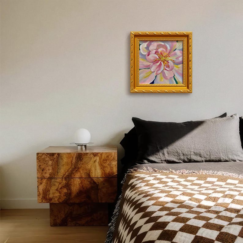 May include: A framed floral painting with a gold ornate frame hangs on a wall. The painting features a pink and yellow flower. A wooden nightstand with a white globe lamp sits below the painting. A bed with a checkered blanket is in the foreground.