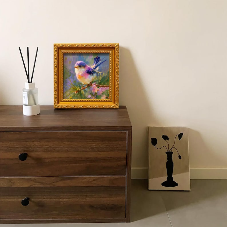 Puede incluir: Una pintura enmarcada de un p&aacute;jaro colorido con tonos azules, morados y naranjas, sobre un fondo de follaje verde y rosa. La pintura est&aacute; en un marco dorado ornamentado. Tambi&eacute;n se ven un difusor y una obra de arte en lienzo.
