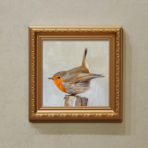 May include: A framed painting of a European robin perched on a wooden post. The bird has a bright orange breast, grey and brown wings, and a long, upright tail. The painting is in a gold ornate frame.