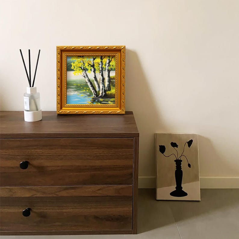 Op de afbeelding: Een ingelijst landschapsschilderij met een gouden, sierlijke lijst, met berkenbomen bij een meer. Een glazen diffuserfles met zwarte rietjes staat op een donkere houten dressoir. Een canvas print met een zwarte vaas en bloemensilhouet.