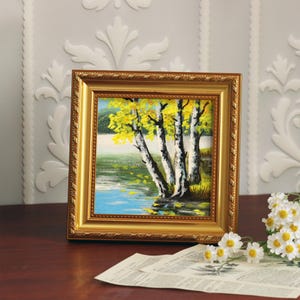 Op de afbeelding: Een ingelijst landschapsschilderij met berken met gele bladeren bij een meer. Het schilderij zit in een gouden, sierlijke lijst. De sc&egrave;ne speelt zich af op een houten oppervlak met bloemen en een krant.