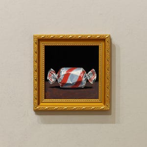 May include: A framed painting of a wrapped sweet with red and white stripes. The sweet is on a brown surface against a black background. The painting is in a gold ornate frame.