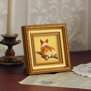 Pu&ograve; includere: Una cornice dorata con un'immagine di un cane corgi visto da dietro, su sfondo giallo. La cornice &egrave; decorata con dettagli ornamentali.