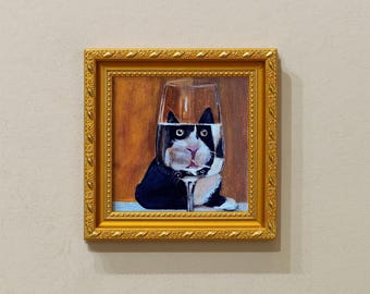 Op de afbeelding: Een ingelijst schilderij van een zwart-witte kat in een wijnglas. Het gezicht van de kat is zichtbaar, met zijn kop en oren in het glas. Het schilderij zit in een gouden lijst en hangt aan een beige muur.