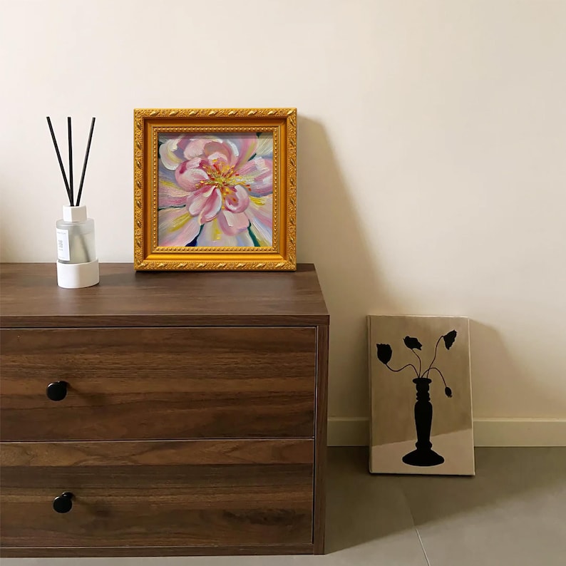 May include: A framed floral painting with a gold ornate frame sits on a dark wood dresser. The painting features a close-up of a pink and yellow flower. A diffuser with black reeds and a vase with a black floral design are also present.