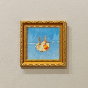 Könnte beinhalten: Gerahmtes Gemälde eines Spiegeleis, das mit zwei Wäscheklammern an einer Wäscheleine vor blauem Himmel hängt. Das Gemälde befindet sich in einem goldenen, verzierten Rahmen. Das Ei hat ein leuchtend orangefarbenes Eigelb.