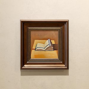 May include: A framed painting shows an open book on a yellow table. The book's pages are visible, with blue and white stripes. The painting has a brown wooden frame with a beaded border, set against a neutral wall.