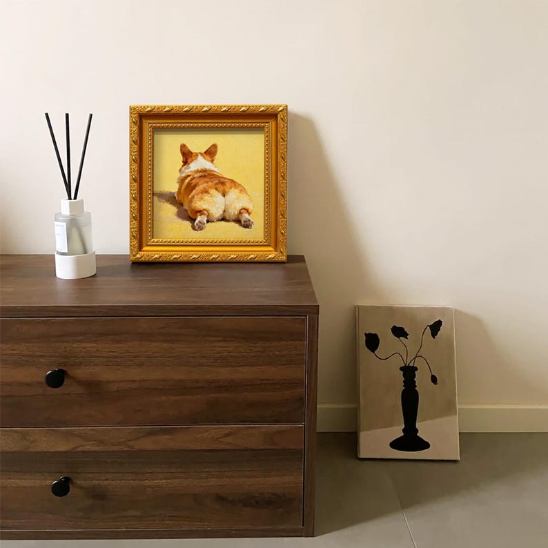 Pu&ograve; includere: Una stampa incorniciata in oro del posteriore di un cane corgi, appoggiata su una cassettiera in legno scuro. La cornice &egrave; dorata con dettagli decorativi. Un diffusore con bastoncini neri e un'opera d'arte minimalista sono presenti.