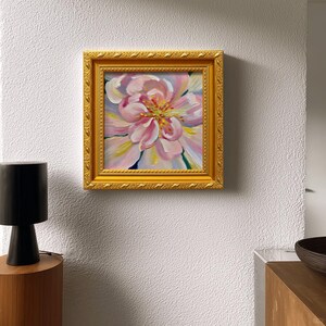 May include: A square framed painting of a pink flower with yellow and white accents. The painting is in a gold ornate frame. The artwork is displayed on a white textured wall. A black lamp and wooden table are in the foreground.