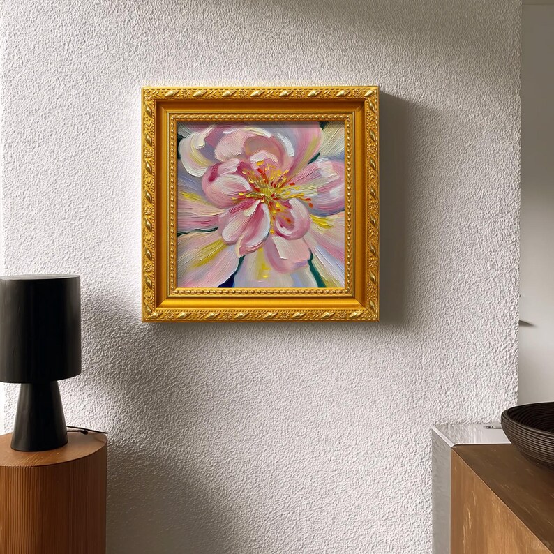May include: A square framed painting of a pink flower with yellow and white accents. The painting is in a gold ornate frame. The artwork is displayed on a white textured wall. A black lamp and wooden table are in the foreground.