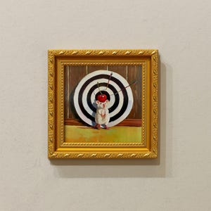 May include: A framed painting featuring a white mouse with a cherry on its head, standing before a target with arrows. The target is black and white, and the painting has a gold frame. The background is a wooden wall.