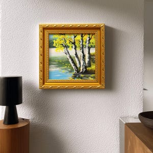 Op de afbeelding: Een vierkant ingelijst schilderij met een gouden, sierlijke lijst. Het kunstwerk toont een landschap met berken met gele bladeren bij een water. Het schilderij is gemonteerd op een witte, getextureerde muur.
