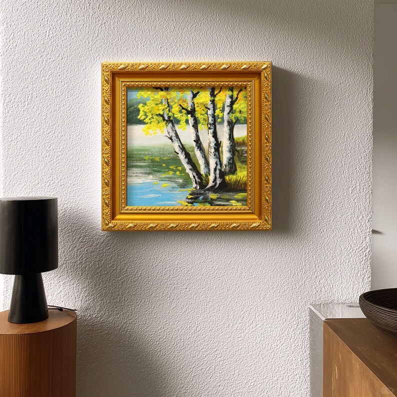 Op de afbeelding: Een vierkant ingelijst schilderij met een gouden, sierlijke lijst. Het kunstwerk toont een landschap met berken met gele bladeren bij een water. Het schilderij is gemonteerd op een witte, getextureerde muur.