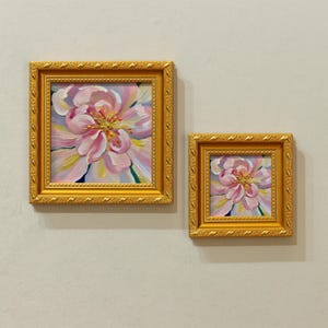 May include: Two framed floral paintings. The larger painting features a close-up of a pink and yellow flower with a detailed center. The smaller painting mirrors the larger one. Both are in ornate gold frames.