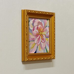 May include: A framed oil painting of a pink flower with yellow and white accents. The artwork is set in a gold-colored ornate frame, creating a classic and elegant aesthetic. The painting is a close-up of the flower's petals.