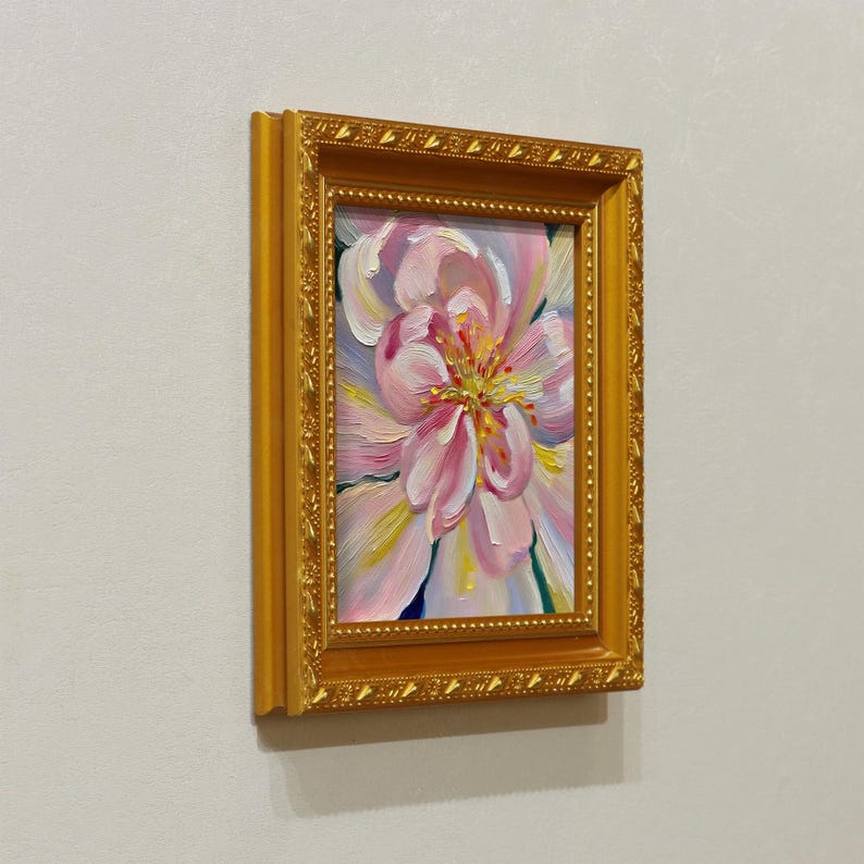 May include: A framed oil painting of a pink flower with yellow and white accents. The artwork is set in a gold-colored ornate frame, creating a classic and elegant aesthetic. The painting is a close-up of the flower's petals.