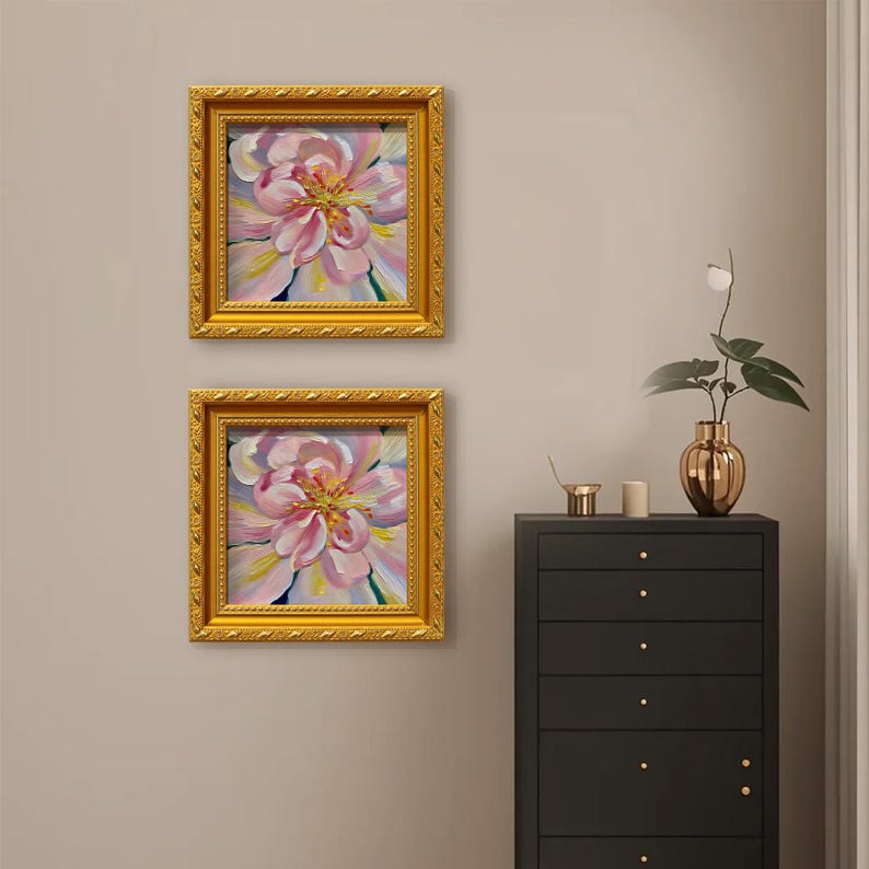 May include: Two framed floral paintings with gold ornate frames. The paintings depict a close-up of a pink flower with yellow and white accents. The paintings are hanging on a neutral wall above a black dresser.