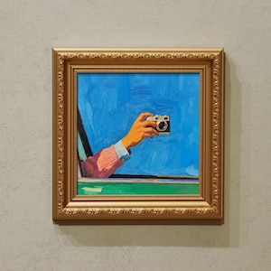 Può includere: Dipinto incorniciato raffigurante una mano che tiene una macchina fotografica da una finestra. Il dipinto ha una cornice dorata, un cielo blu e una linea di orizzonte verde. La mano è arancione e la manica rosa.