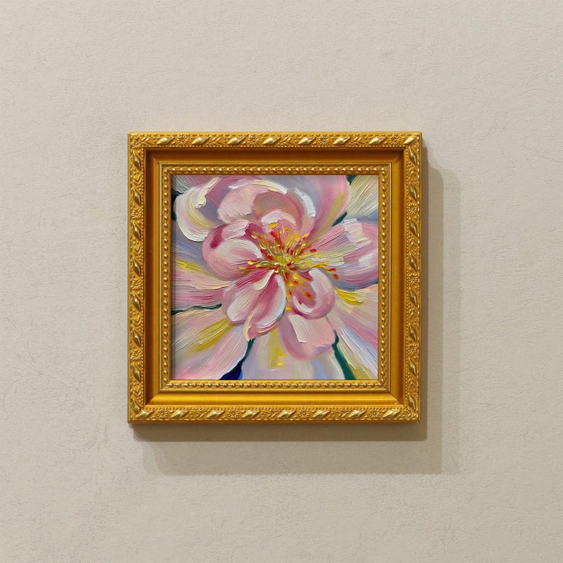 May include: A framed floral painting featuring a large, pink flower with yellow and white accents. The artwork is set within a gold-colored, ornate frame, creating a classic and elegant aesthetic. The painting is mounted on a neutral-toned wall.