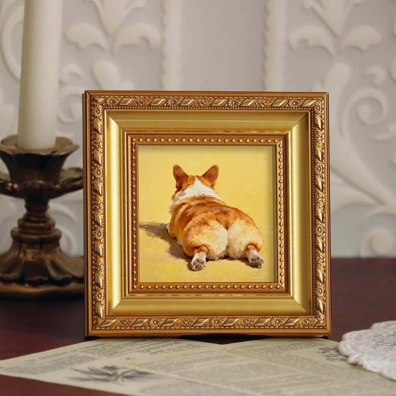 Pu&ograve; includere: Dipinto incorniciato di un cane corgi, visto da dietro, appoggiato su uno sfondo giallo. Il cane ha pelo marrone e bianco. La cornice dorata presenta dettagli ornati. Il dipinto &egrave; esposto su una superficie scura.