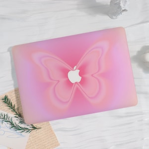 Könnte beinhalten: Rosa Laptop-Hülle mit Schmetterlingsdesign. Die Hülle hat einen Farbverlauf, der von einem helleren Rosa zu einem tieferen Farbton übergeht. Das Apple-Logo ist in der Mitte des Schmetterlingsdesigns sichtbar. Die Hülle liegt auf einer weißen Holzoberfläche.