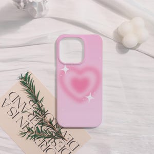 May include: A light pink phone case featuring a gradient heart design in shades of pink. The case has a rectangular shape with rounded corners and a cutout for the camera. Two white starbursts are on the case. The case is on a white surface.