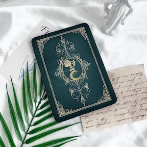 May include: A green journal with a gold design featuring a crescent moon and a rose. The journal has a decorative border and a vintage aesthetic.