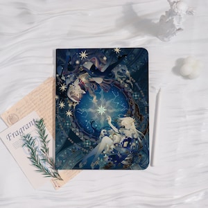 May include: A dark blue notebook with an illustration of a celestial scene featuring figures, stars, and a central starburst design. The word "Fragrant" is visible on a card next to the notebook. A white stylus and decorative items are also present.