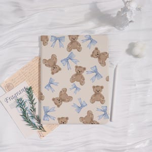 May include: A beige tablet case featuring a pattern of brown teddy bears and light blue bows. The case has a smooth surface and a rectangular shape. A white stylus is placed on the right side of the case.