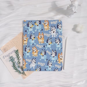 May include: A light blue notebook featuring a repeating pattern of cartoon dogs in various poses. The word "BLUEY" is printed in white on the notebook. The notebook is open, revealing blank pages.