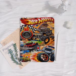 May include: A colorful Hot Wheels-themed tablet case featuring monster trucks and flames. The design includes the Hot Wheels logo and various monster truck illustrations in vibrant colors. The case is rectangular and likely made of a durable material.