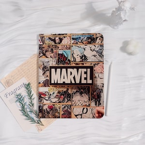 Könnte beinhalten: Ein Notizbuch im Marvel-Design mit einem Comic-Panel-Collage-Cover. Das MARVEL-Logo in Schwarz ist prominent platziert. Das Notizbuch liegt zusammen mit Schreibwaren und dekorativen Gegenständen auf einer weißen Oberfläche.