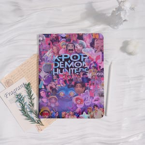 May include: A rectangular notebook with a collage of colorful cartoon characters and the text "KPOP DEMON HUNTERS" in blue and purple. The notebook is surrounded by decorative elements, including a small statue and a sprig of greenery.