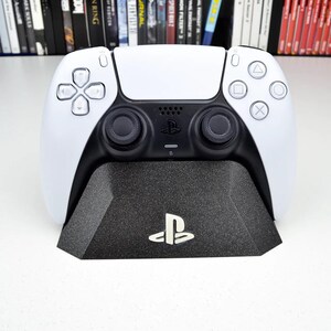 Op de afbeelding: Een wit en zwarte PlayStation 5-controller op een zwart oplaadstation. De controller heeft een symmetrisch ontwerp met knoppen en joysticks. De standaard heeft het PlayStation-logo.