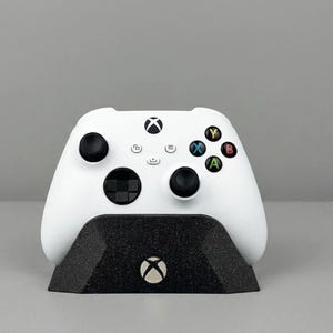 May include: A white Xbox Series X/S controller resting on a black textured charging stand. The controller has black buttons and colorful action buttons. The Xbox logo is visible on both the controller and the stand.