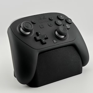 Pode incluir: Um comando Nintendo Switch Pro preto com uma pega texturizada, botões, joysticks e uma cruz direcional. O comando está sobre um suporte preto. O logótipo Nintendo Switch é visível.