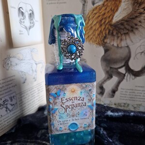 Può includere: Una bottiglia di vetro blu piena di un liquido blu scintillante e un ciondolo d'argento con una pietra turchese. La bottiglia ha un'etichetta che recita "Essenza di Spepanza" e "Un sorso e la luce tornera a brillare."