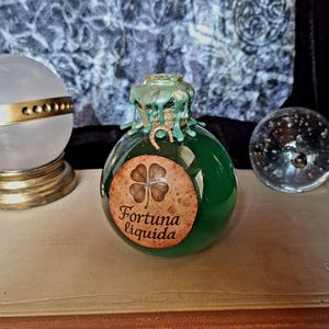 Può includere: Una bottiglia rotonda di vetro verde con un sigillo di cera e un'etichetta che recita "Fortuna Liquida" con un quadrifoglio. La bottiglia è su un libro. Una sfera di vetro e una lampada decorativa sono sullo sfondo.