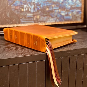 Puede incluir: Un libro encuadernado en cuero marrón con varios marcadores de cinta. El libro está cerrado y apoyado sobre una superficie de madera oscura.