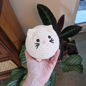 May include: A handmade, cream-coloured crochet cat toy with black embroidered eyes and whiskers, and a pink nose. The cat is round and plush, and is being held in a hand. The background includes green plants and a wooden cabinet.