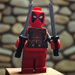 Puede incluir: Una figura Lego roja de Deadpool, sosteniendo dos katanas plateadas. La figura tiene una máscara en blanco y negro y un diseño de torso detallado. Está sobre un pequeño bloque de madera marrón, con un fondo de piedra.