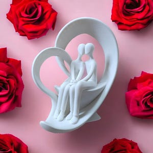 Könnte beinhalten: Weiße Keramikskulptur von zwei Figuren, die auf einem herzförmigen Sockel sitzen. Die Skulptur befindet sich auf einem rosa Hintergrund mit roten Rosen.