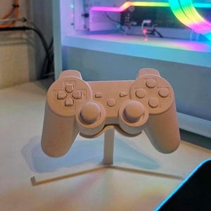 Könnte beinhalten: Ein weißer Plastik-Videospielcontroller auf einem weißen Ständer. Der Controller hat ein Standardlayout mit Knöpfen, Joysticks und einem Steuerkreuz.