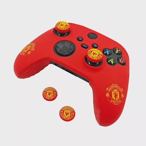 May include: A red gaming controller with yellow and red Manchester United branding. The controller has black buttons and a directional pad. Includes additional thumb grips with the same logo.